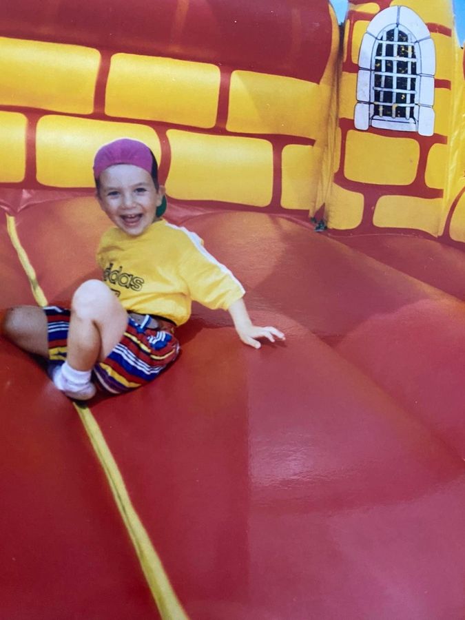Eftim de niño, con gorra rosa y camiseta amarilla, riendo sobre un castillo hinchable rojo