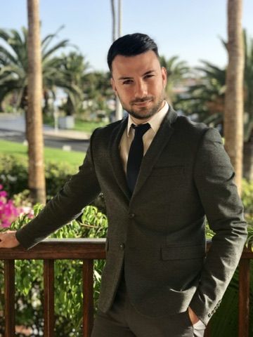 Eftim con traje oscuro y corbata, apoyado en una barandilla de madera entre palmeras